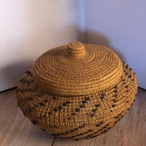 May include: A round, lidded basket made of woven natural fibers. The basket has a textured surface with a repeating pattern of dark brown accents near the base. The lid features a central knob.