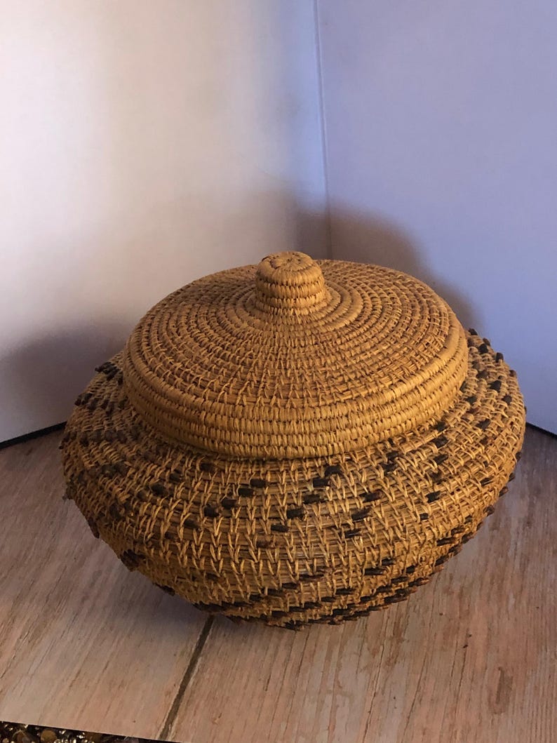 May include: A round, lidded basket made of woven natural fibers. The basket has a textured surface with a repeating pattern of dark brown accents near the base. The lid features a central knob.