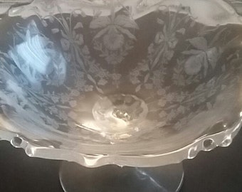 May include: A clear glass pedestal bowl with a floral etched design. The bowl has a scalloped rim and a round base. The etched design features flowers and foliage. The bowl is likely used for serving or display.