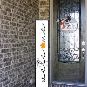 May include: A tall, rectangular welcome sign with a black frame and white background. The word "welcome" is written in a cursive font, with an orange leaf graphic. The sign is placed on a porch next to a brick wall.