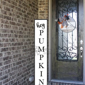 Può includere: Un cartello verticale bianco con la scritta nera "hey PUMPKIN" è esposto accanto a un muro di mattoni. Una ghirlanda con un fiocco e dettagli arancioni è appesa alla porta. Un tappetino di benvenuto è alla base della porta.