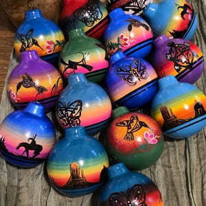 May include: A collection of hand-painted gourd ornaments. Each ornament features a unique design, including butterflies, hummingbirds, and desert landscapes, set against a gradient of colors. The ornaments have a loop for hanging.