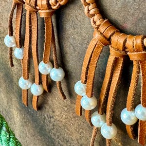 Puede incluir: Un collar de cuero marrón con detalles de cuero trenzado y perlas. El collar tiene múltiples hebras de cuero con borlas y perlas.