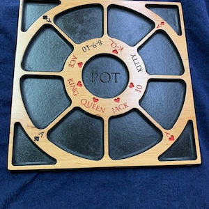 May include: A wooden game board with a circular center labeled "POT" and eight surrounding sections labeled "Ace", "King", "Queen", "Jack", "Kitty", "10", "01-6-8", and "7-K". Each section has a small red heart symbol and a playing card symbol.