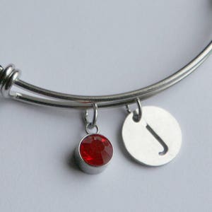 May include: A silver bangle bracelet with a red crystal charm and a silver charm with the letter "J" engraved on it.