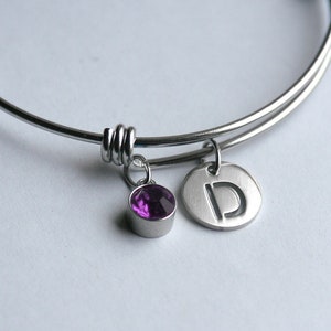 May include: A silver bangle bracelet with a purple crystal charm and a silver charm with the letter "D" engraved on it.
