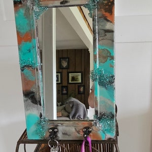 May include: A turquoise, brown, and black resin mirror with a decorative glass bead border. The mirror has two hooks on the bottom for hanging keys or other items.