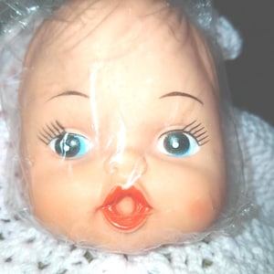 May include: A vintage doll head with blue eyes, long eyelashes, and red lips, encased in plastic. The doll head is set against a white crocheted background, suggesting a handmade or retro aesthetic. The doll's skin is a pale peach color.