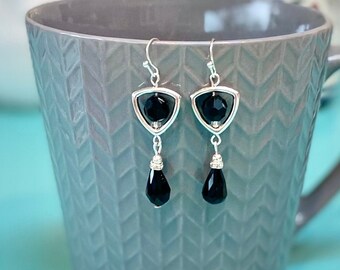 May include: A pair of silver earrings with black teardrop-shaped crystals. The earrings feature a triangular frame with a smaller black crystal in the centre.