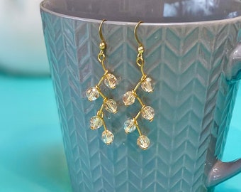 May include: A pair of gold dangle earrings with clear crystal beads. The earrings are hanging from a grey ceramic mug.