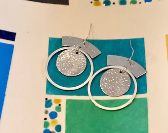 May include: A pair of silver earrings featuring a textured circle and a curved bar design. The earrings are shown against a colourful tiled background.