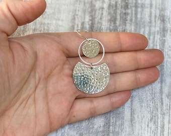 May include: Silver earrings with a hammered texture. The earrings have a small circle and a larger, half-moon shape, connected by a small ring. The earrings are displayed against a light-coloured background.