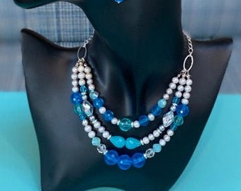 May include: A three-strand necklace and matching earrings set. The necklace features blue, clear, and white beads. The earrings have a blue and a green bead. The jewellery is displayed on a black mannequin head.