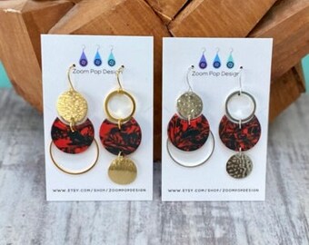 May include: Two pairs of gold and silver earrings featuring red and black patterned circles and hammered metal accents. The earrings are displayed on white cards with the text "Zoom Pop Design" and a website address.