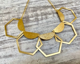 May include: A gold necklace with a chain and geometric pendants. The design includes open hexagons and half-circle shapes, creating a modern accessory. The necklace is displayed on a light-coloured surface.