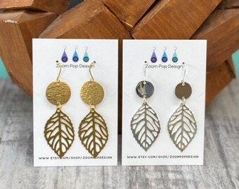 May include: Two pairs of earrings, one gold and one silver, with a leaf design and a round disc. The earrings are displayed on a white card with the text "Zoom Pop Design" and a website address.