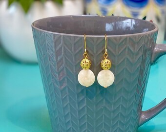 May include: A pair of gold dangle earrings with white round beads. The earrings are hanging from a grey ceramic mug.