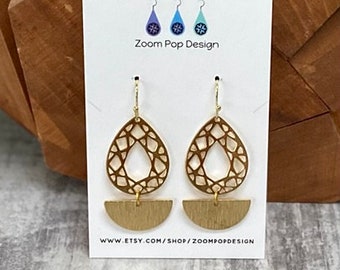 May include: Gold tone earrings with a geometric design. The earrings feature a teardrop-shaped top with a cut-out pattern and a half-circle bottom. The earrings are on a white card with the text "Zoom Pop Design" and a website address.