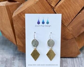 May include: Silver and gold earrings with a round and diamond shape. The earrings are hanging from a white card with the text "Zoom Pop Design" and a website address.