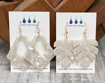 May include: Two pairs of white earrings with gold hooks. One pair is a geometric shape with a cutout, and the other pair is a leaf shape.