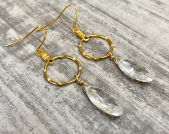 May include: Gold dangle earrings with a clear teardrop crystal hanging from a textured gold circle.