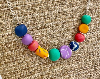 May include: A colourful necklace featuring round and cylindrical beads in shades of blue, purple, pink, orange, green, and yellow. The beads are strung on a silver chain.