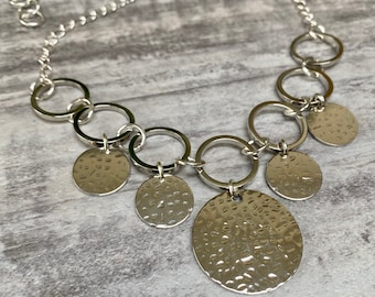 May include: A silver necklace featuring a chain and several hammered metal circles. The circles are of different sizes and are attached to the chain.