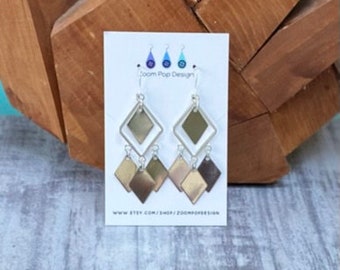 May include: Silver dangle earrings with multiple layered diamond-shaped charms. The charms are silver and gold in colour. The earrings are displayed on a white card with the text "Zoom Pop Design" and a website address.