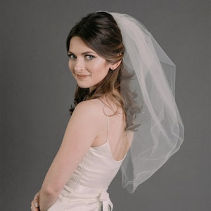 Op de afbeelding: Een vrouw in een witte satijnen trouwjurk met een simpel, elegant ontwerp. Ze draagt een eenlaags tule sluier met een delicate parelrand. De sluier is over haar hoofd en schouders gedrapeerd, en ze kijkt over haar schouder met een zachte glimlach.