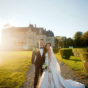 May include: A bride and groom pose for a wedding portrait in front of a large, white mansion. The bride is wearing a white strapless gown with a long train and a veil. The groom is wearing a black tuxedo with a white bow tie. The couple is standing on a path in front of a lush green lawn.