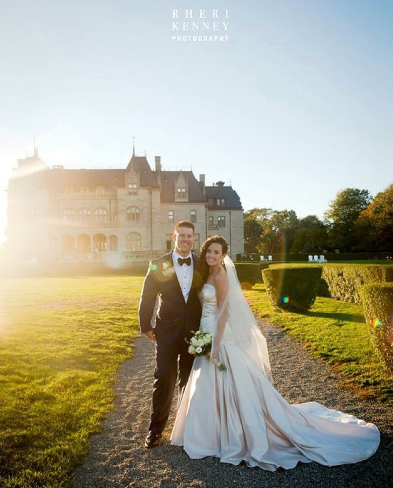 May include: A bride and groom pose for a wedding portrait in front of a large, white mansion. The bride is wearing a white strapless gown with a long train and a veil. The groom is wearing a black tuxedo with a white bow tie. The couple is standing on a path in front of a lush green lawn.
