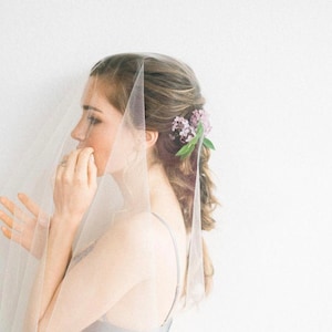 May include: A woman wearing a white veil with a floral hairpiece. The veil is sheer and covers her face. She is looking to the side.