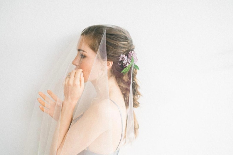 May include: A woman wearing a white veil with a floral hairpiece. The veil is sheer and covers her face. She is looking to the side.