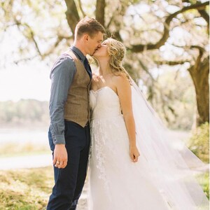 May include: A bride and groom kiss under a large tree with green foliage. The bride is wearing a white strapless wedding dress with a veil. The groom is wearing a blue button-down shirt, a brown vest, and dark blue pants.