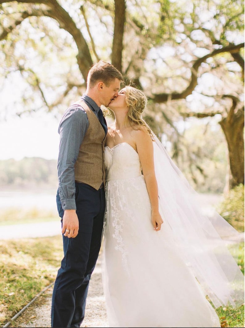 May include: A bride and groom kiss under a large tree with green foliage. The bride is wearing a white strapless wedding dress with a veil. The groom is wearing a blue button-down shirt, a brown vest, and dark blue pants.