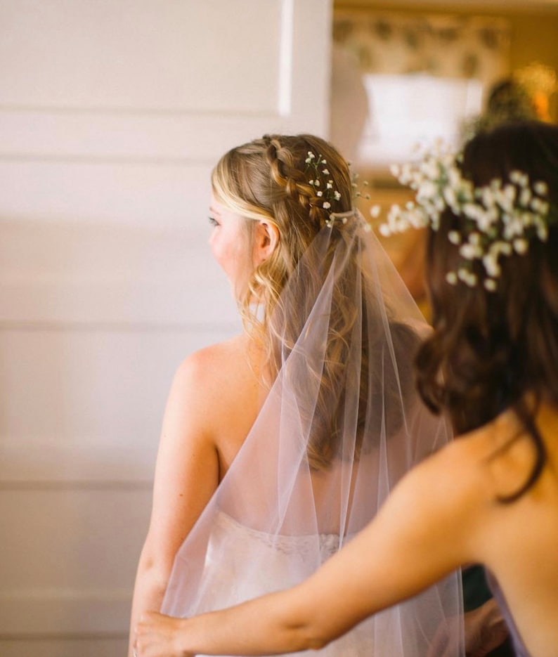 May include: A bride with a white veil and a floral crown is having her veil adjusted by a bridesmaid.