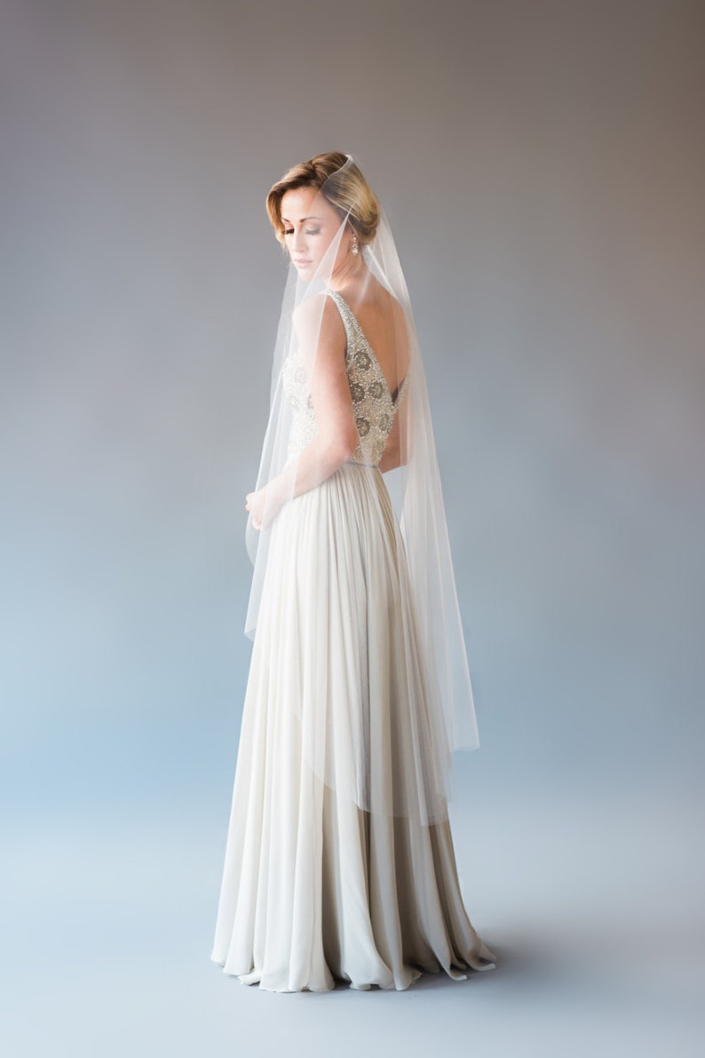 May include: A bride wearing a white wedding dress with a beaded bodice and a long veil. The dress has a flowing skirt and the bride is looking down.
