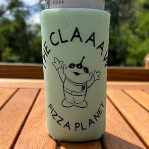 Puede incluir: Un enfriador de latas verde con el texto "The Claaaw Pizza Planet" y un extraterrestre de dibujos animados con tres ojos y dos brazos levantados en el aire.