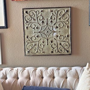 May include: Square, ornate metal wall art with a light green and brown finish. The design features scrolling vines and floral accents within a dark brown frame. The artwork is mounted on a beige wall above a tufted sofa.