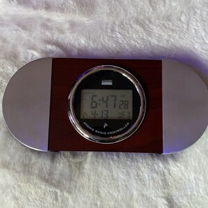 May include: A silver and wood framed digital clock with a black face. The clock displays the time 6:47, the date 12/28, and the day of the week WE. The clock is labeled "Atomic Radio Controlled".