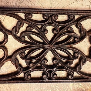 May include: A rectangular, cast iron decorative mat with an ornate, symmetrical scrollwork design. The mat is dark brown with a weathered appearance, and the openwork allows the background to show through.