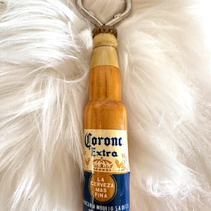 May include: A bottle opener shaped like a Corona Extra beer bottle. The opener has a silver metal top and a wooden body with the Corona Extra logo and text in blue and gold. The bottle opener is on a white, fluffy surface.