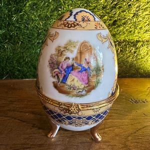 May include: An egg-shaped decorative trinket box with a hinged lid. The white porcelain box features a gold trim, blue and gold patterned base, and a painted scene of a couple. The box is supported by three gold feet.