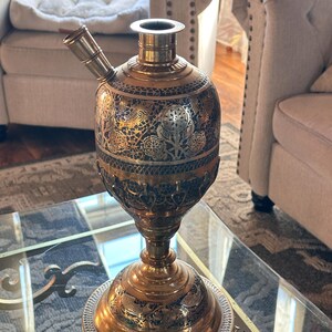 May include: Ornate brass water pipe with intricate etched designs. The vessel features a bulbous body, a flared base, and a curved mouthpiece. The brass has a warm, golden hue, with darker accents in the etched patterns. The piece is displayed on a glass-topped table.