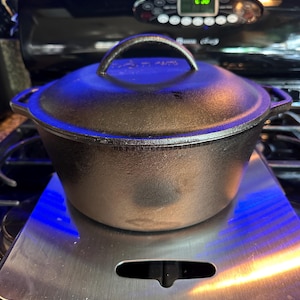 May include: A black cast iron Dutch oven with a lid and handle. The pot is sitting on a stainless steel surface. The oven is on a black stove top. The handle on the lid is arched.