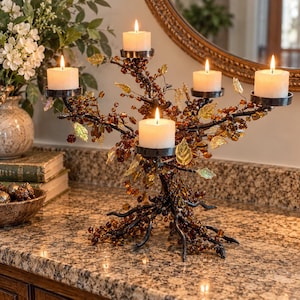 May include: A five-arm candelabra featuring a dark, branch-like metal structure adorned with amber-colored beads and translucent leaf accents. Five white pillar candles are placed in circular metal holders atop the branches.