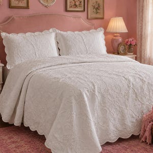 May include: A white bedspread and two matching pillow shams featuring an intricate, raised floral and scrollwork pattern. The bedding set has scalloped edges and a textured, quilted finish that covers the entire surface of the bed.