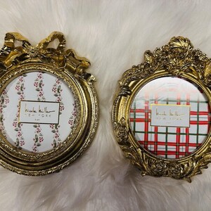 May include: Two ornate, gold-colored picture frames. One has a bow design at the top, the other has a floral design. Both have a round opening with a patterned background and a "Nicole Miller New York" label.