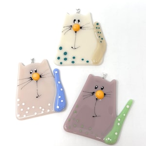 May include: Three whimsical cat pendants in cream, mauve, and lavender. Each cat has a round orange nose, black whiskers, and a tail with polka dots. The cream cat has teal dots, the mauve cat has blue dots, and the lavender cat has green dots.