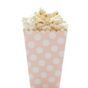 May include: A pink popcorn box with white polka dots is filled with fluffy, white popcorn. The box has a scalloped top edge and is set against a white background. The popcorn is overflowing from the top of the box.